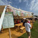 Outdoor event with people gathered around tables under a large tent with colourful ribbons, featuring a 'Latitude' menu board.
