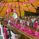 Decorated indoor event space with pink and yellow decorations, including a long wooden table with pink runners and chairs.