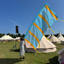 Person standing next to a large blue flag with orange stripes in an outdoor setting with tents
