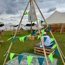 Person sitting on a wooden crate under a decorative teepee with green bunting at a festival